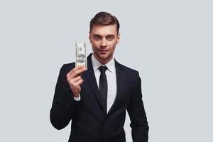 mått av Framgång. Bra ser ung man i full kostym leende och innehav en papper valuta medan stående mot grå bakgrund foto