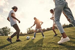 lekfull vänner. grupp av ung leende människor i tillfällig ha på sig spelar fotboll medan stående utomhus foto