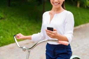 Bra Nyheter från vänner. närbild av ung kvinna innehav mobil telefon och leende medan gående med henne cykel i parkera foto