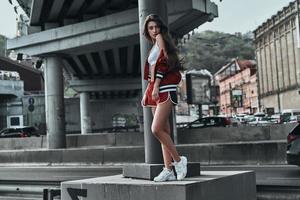 stad stil. full längd av skön ung kvinna i sport Kläder ser över axel medan Framställ under de bro utomhus foto