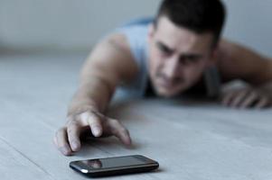 hjälp mig frustrerad ung man liggande på de golv och stretching hand till de mobil telefon foto