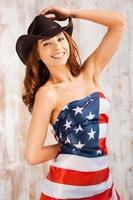 stolt amerikansk. skön ung bar överkropp cowgirl justeras henne hatt och beläggning själv förbi amerikan flagga medan stående mot de trä- bakgrund foto