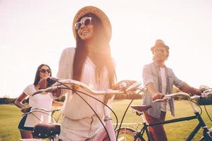 njuter en bra sommar dag. glad ung människor tryckande deras cyklar medan gående förbi grön gräs utomhus foto