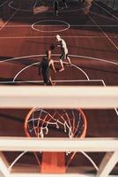 två ung män i sporter Kläder spelar basketboll och leende medan utgifterna tid utomhus foto