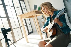 glad ung kvinna leende och spelar gitarr medan spelar gitarr i främre av digital kamera foto