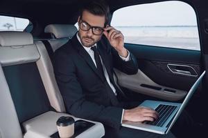 planera hans företag. omtänksam ung man i full kostym arbetssätt använder sig av bärbar dator och justeras hans glasögon medan Sammanträde i de bil foto
