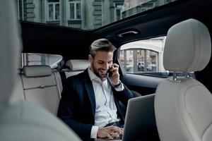 diskuterar företag detaljer. stilig ung man i full kostym talande på smart telefon och leende medan Sammanträde i de bil foto