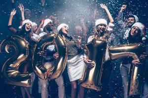 önskar en Lycklig ny år. grupp av glad ung människor i santa hattar bärande guld färgad tal och kasta konfetti foto