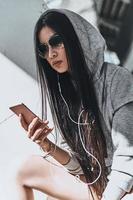 musik i henne hjärta. skön ung kvinna i tillfällig ha på sig innehav en smart telefon medan Sammanträde utomhus foto