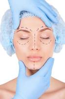förberedelse för ansiktsbehandling kirurgi. attraktiv ung kvinna i medicinsk huvudbonader och skisser på ansikte förvaring ögon stängd medan läkare granskning henne ansikte isolerat på vit foto