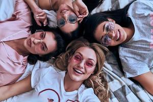 oemotståndligt flickor. topp se av fyra lekfull ung kvinnor i glasögon leende och ser på kamera medan liggande på de säng på Hem foto