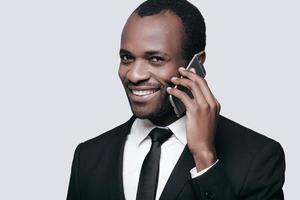 känsla Lycklig. stilig ung afrikansk man leende och talande på de telefon medan stående mot grå bakgrund foto