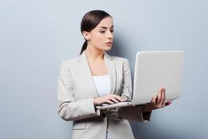 upptagen med henne arbete. självsäker ung affärskvinna innehav bärbar dator medan stående mot grå bakgrund foto