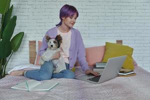 självsäker Tonårs flicka använder sig av bärbar dator medan Sammanträde på säng med henne hund foto