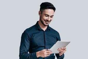 använder sig av modern teknik. stilig ung man i skjorta ser på hans digital läsplatta och leende medan stående mot grå bakgrund foto