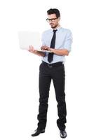 stödjande din företag. full längd av stilig ung man i skjorta och slips arbetssätt på bärbar dator medan stående mot vit bakgrund foto