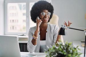 skön ung afrikansk kvinna talande på mobil telefon och leende medan Sammanträde på henne arbetssätt plats i kontor foto