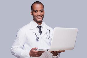 självsäker medicinsk läkare. självsäker afrikansk läkare innehav bärbar dator och leende medan stående mot grå bakgrund foto