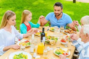 bön- innan middag. topp se av familj av fem människor innehav händer och bön- innan middag medan Sammanträde på de tabell utomhus foto