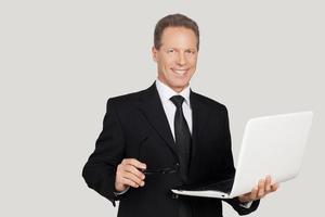 framställning din företag lätt. glad senior man i formell klädsel innehav bärbar dator och leende medan stående mot grå bakgrund foto
