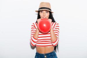 baloon roligt. lekfull ung kvinna blåser röd ballong medan stående mot vit bakgrund foto
