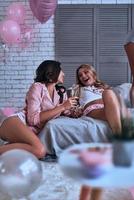 underbar skönheter. två skön ung kvinnor i natt ha på sig talande och leende medan dricka champagne i de sovrum foto