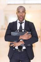 frustrerad affärsman. chockade ung afrikansk man i full kostym innehav en väska i hans händer och förvaring mun öppen medan ser på kamera foto