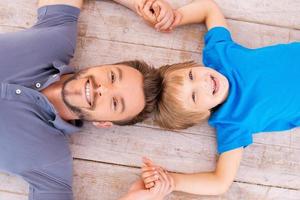 Lycklig pappa och son. topp se av Lycklig far och son innehav händer leende på kamera medan liggande på de hårt träslag golv foto