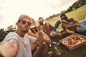 själv porträtt av ung människor i tillfällig ha på sig leende medan njuter pizza och öl utomhus foto