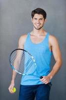 redo till spela. leende ung muskulös man innehav tennis racket medan stående mot grå bakgrunder foto