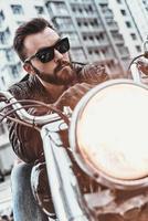 verklig macho. stilig ung man i läder jacka och solglasögon ridning motorcykel medan utgifterna tid utomhus foto