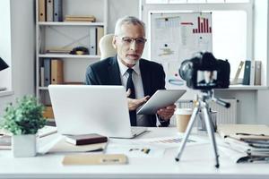 senior man i elegant företag kostym använder sig av digital läsplatta medan framställning social media video foto
