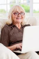surfing de netto är roligt. senior kvinna arbetssätt på bärbar dator och leende medan Sammanträde på de stol foto