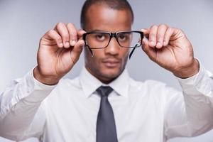 tid för ny glasögon. självsäker ung afrikansk man i skjorta och slips innehav glasögon och ser genom dem medan stående mot grå bakgrund foto