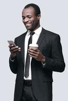 tar emot trevlig meddelanden stilig ung afrikansk man ser på mobil telefon och leende medan stående mot grå bakgrund foto