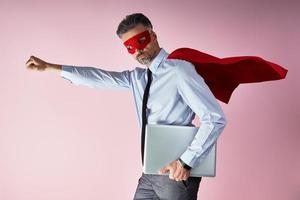 självsäker man i skjorta och slips bär superhjälte cape och bärande bärbar dator mot rosa bakgrund foto
