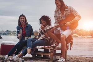 spelar romantisk låt. grupp av ung människor i tillfällig ha på sig leende medan njuter strand fest nära de lägereld foto