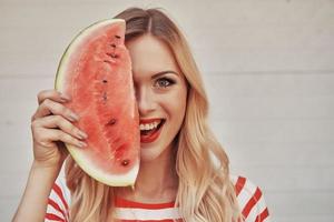 sommar smak. glad ung kvinna innehav skiva av vattenmelon mot halv del av henne ansikte och leende medan stående utomhus foto