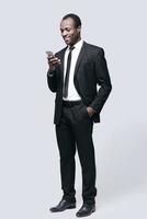 läsning trevlig meddelande full längd av stilig ung afrikansk man ser på mobil telefon och leende medan stående mot grå bakgrund foto