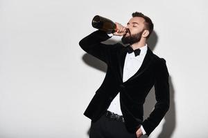fira något särskild. stilig ung man i full kostym dricka champagne från de flaska medan stående mot grå bakgrund foto