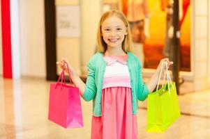 liten shopaholic. glad liten flicka innehav färgrik handla väska och leende medan stående i handla köpcenter foto