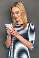 meddelande till vän.vacker ung kvinna använder sig av henne smart telefon och ser på den med leende medan stående mot grå bakgrund foto