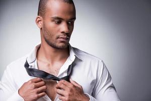 tar hans slips bort. stilig ung svart man tar av hans slips medan stående mot grå bakgrund foto