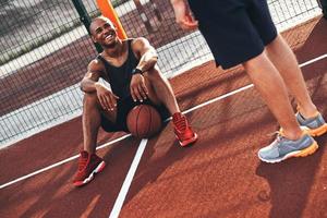 spel är över. ung afrikansk man i sporter Kläder leende medan Sammanträde på de basketboll fält utomhus foto