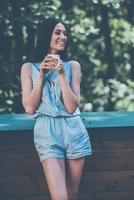 njuter färsk kaffe. skön ung kvinna dricka kaffe och leende medan stående utomhus med grön träd i de bakgrund foto