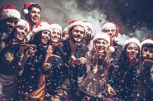 liten jul magi. grupp av skön ung människor i santa hattar blåser färgrik konfetti och ser Lycklig foto