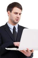 arbetssätt på bärbar dator. självsäker ung man i formell klädsel arbetssätt på bärbar dator medan stående isolerat på vit foto
