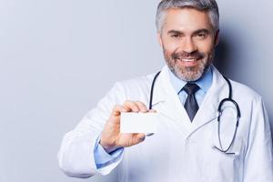 läkare innehav företag kort. glad mogna läkare som visar hans företag kort och leende medan stående mot grå bakgrund foto