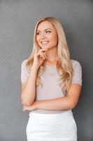 tänkande positiv. Lycklig ung blond hår kvinna innehav hand på haka och ser bort medan stående mot grå bakgrund foto