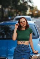 skön flicka sitter på de huva av en blå bil foto